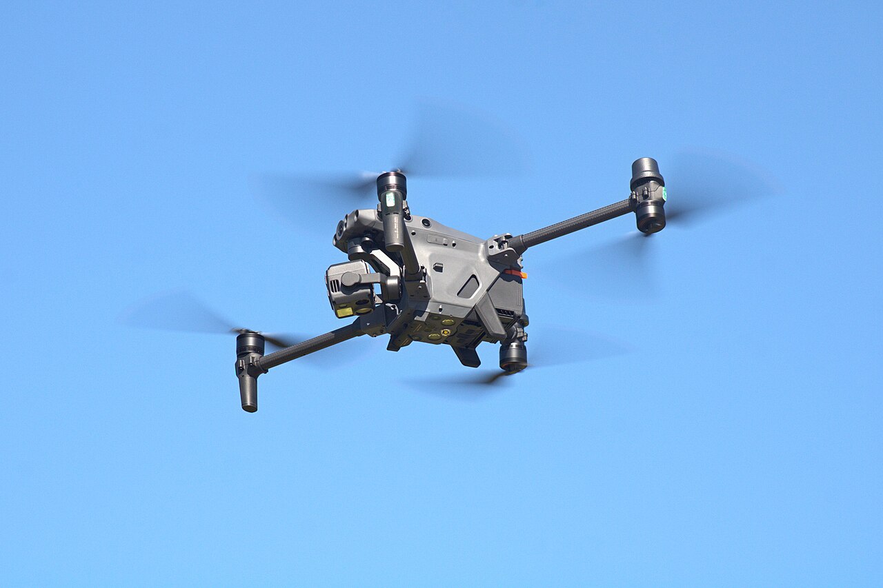 A DJI Matrice 30T inspection quadcopter in flight against a clear blue sky, rotors spinning, with its stabilized camera gimbal mounted under the nose.