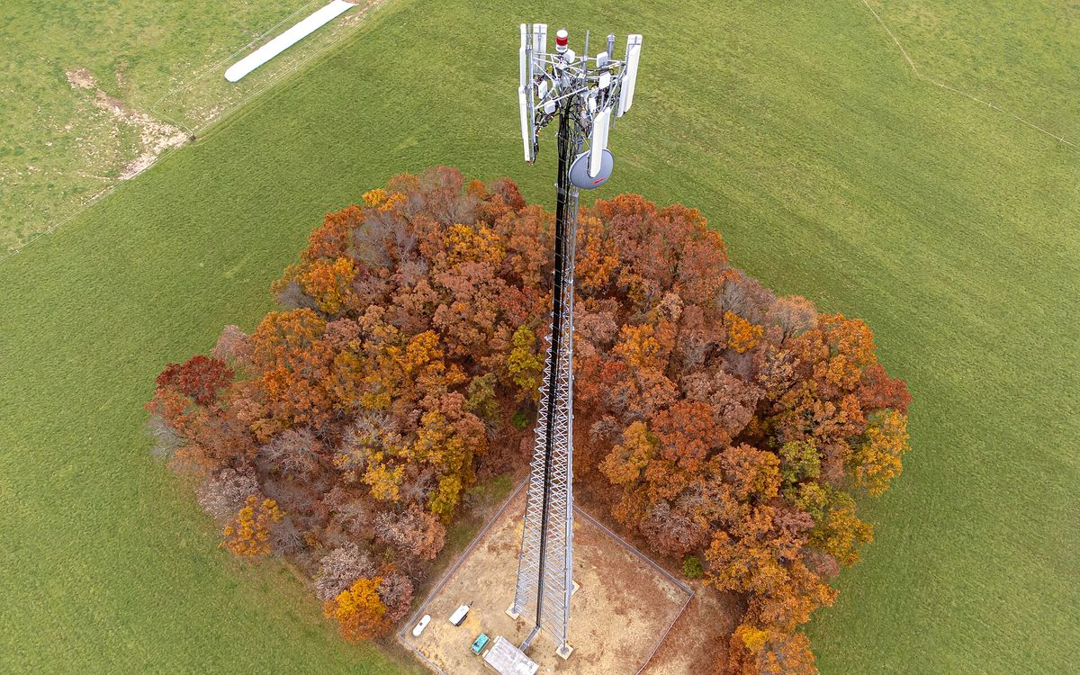 Drone view looking down on a lattice cell tower rising from a ring of autumn trees in a green field